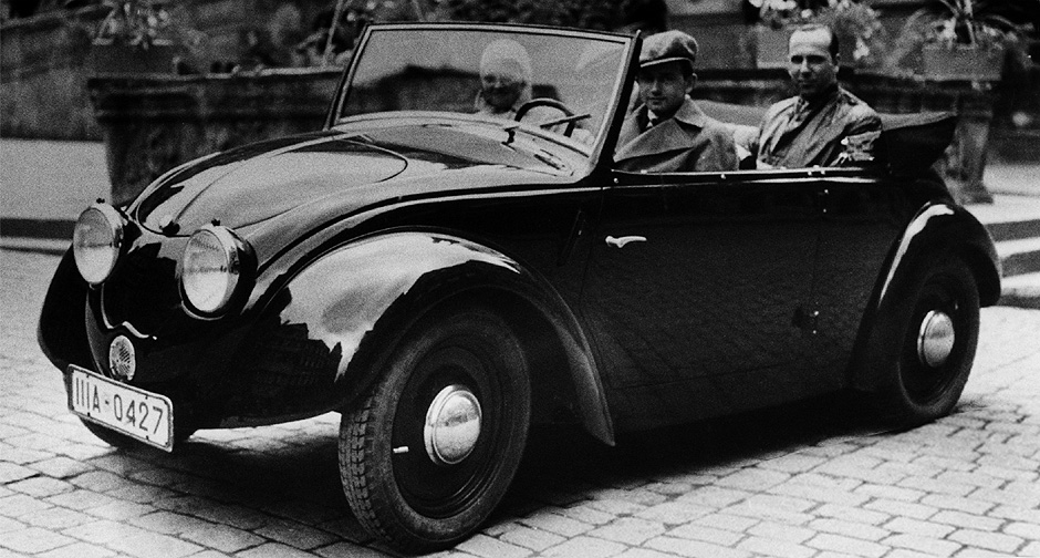 1935. Ferry Porsche at the wheel of the second Volkswagen prototype (V2) on the market square in Tübingen. Beside him is his wife Dorothea and on the backseat is Hellmuth Zarges, a friend of the Porsche family.
