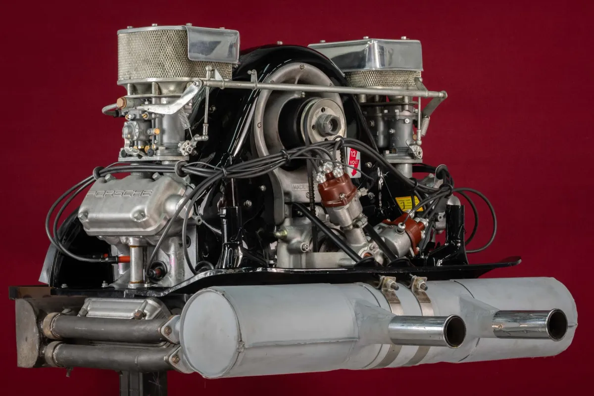 The Type 547 “Fuhrmann” engine that powered the Porsche 550 was a 1,498 cc air-cooled flat four built around an aluminum crankcase with a roller-bearing crankshaft — a choice that reduced friction and allowed sustained high rotational speeds. Each cylinder bank carried dual overhead camshafts driven by vertical shafts and bevel gears, eliminating the timing elasticity associated with chains and providing exceptional valve-event precision at elevated RPM. The hemispherical combustion chambers employed dual spark plugs per cylinder, improving flame propagation and combustion stability under variable load and altitude conditions. Dry-sump lubrication ensured consistent oil delivery during prolonged high-speed cornering and extended flat-out operation, critical for endurance competition. Twin carburetors supplied mixture through short intake tracts designed to optimize volumetric efficiency without sacrificing throttle response. With early outputs around 110 horsepower and later evolutions exceeding 130 horsepower at over 7,000 RPM, the 547 was engineered not merely for peak figures but for mechanical survivability at sustained revs — a defining advantage in long-distance racing.