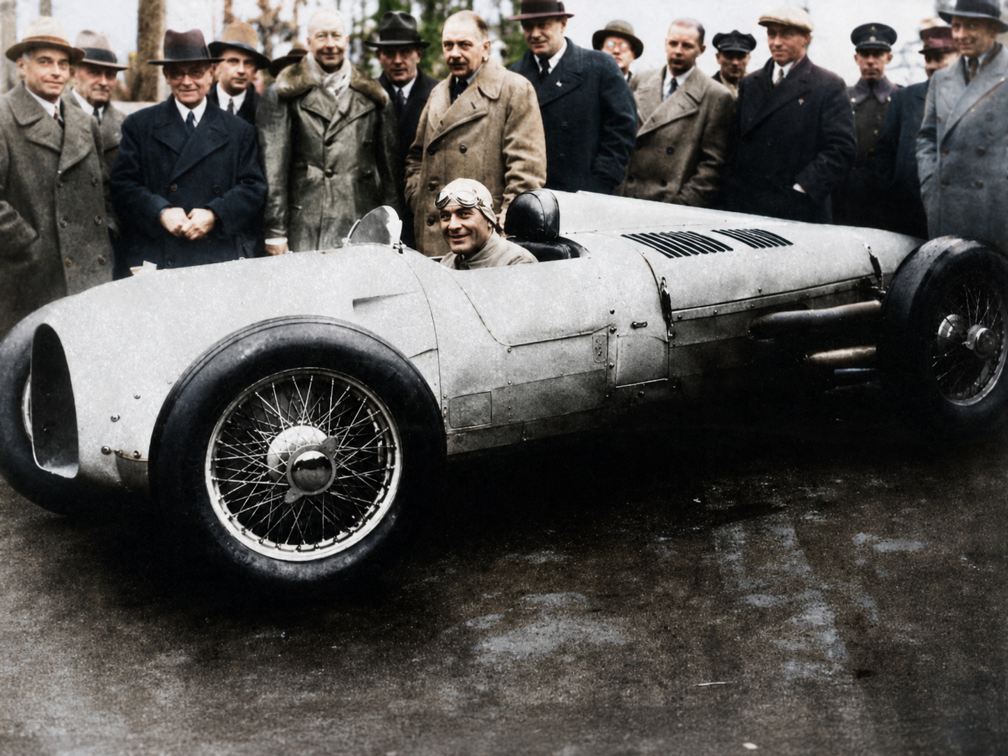 Hans Stuck sits at the wheel of the Auto Union Type A as Ferdinand Porsche and other members of the team gather around the revolutionary new Grand Prix racer. The compact nose and elongated rear body reflected its unconventional architecture, with the supercharged V16 positioned behind the driver rather than ahead of him. The scene captures the moment when Porsche’s speculative P-Wagen concept became a working competition car—and one of the most closely watched machines in European motorsport.