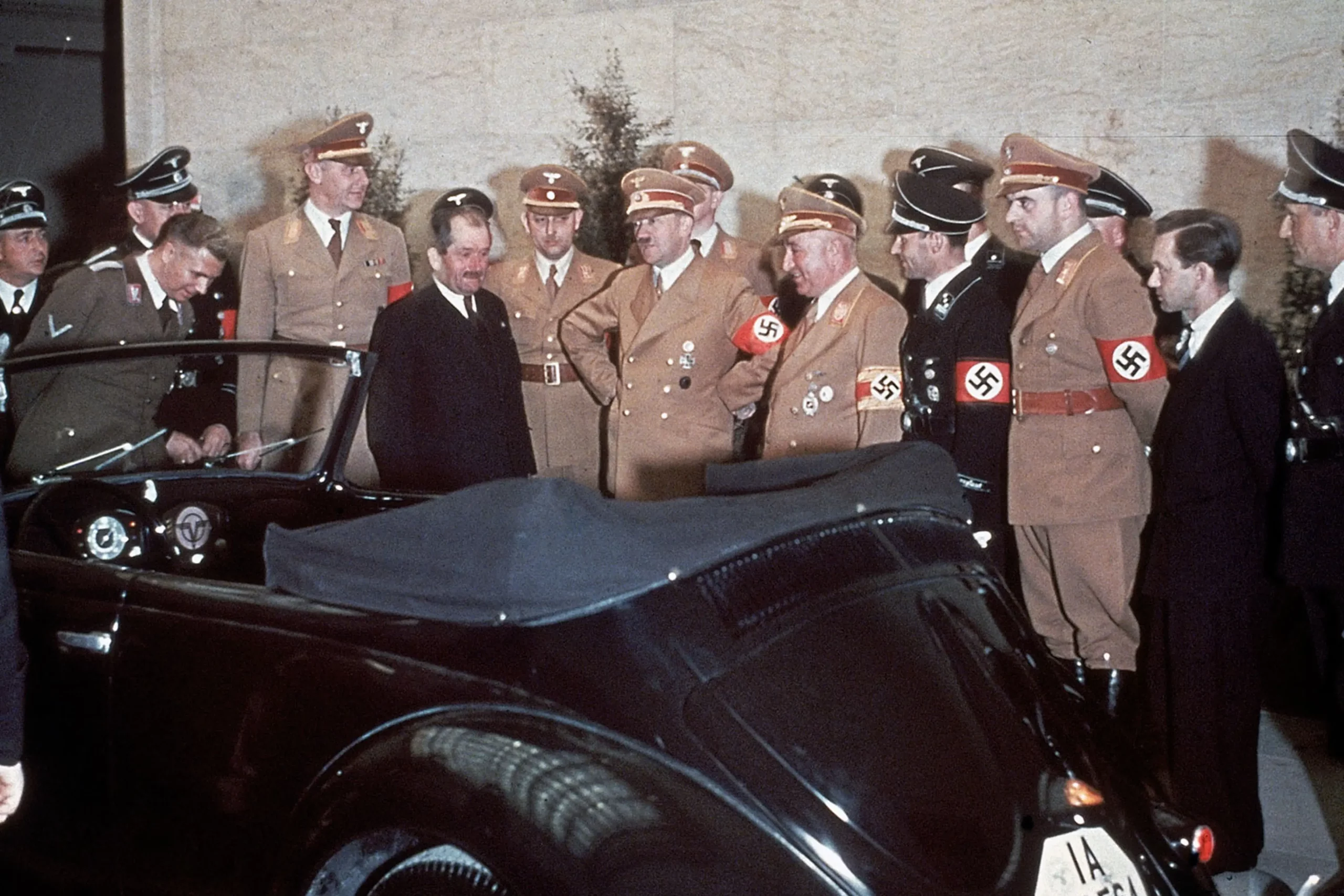 Presented on April 20, 1939, as a gift marking Adolf Hitler’s fiftieth birthday, this specially prepared convertible Volkswagen became a carefully staged symbol of the regime’s claim that political power, industrial planning, and modern engineering were moving in the same direction. Ferdinand Porsche, standing in the dark suit, appears beside Hitler and Robert Ley, head of the German Labour Front, as the group inspects the car before an audience of uniformed officials. The scene captures Porsche not simply as the designer of a new automobile, but as a visible participant in the ceremonial world surrounding Nazi leadership, where engineering achievements were folded directly into propaganda and personal loyalty to the Führer. Appearances such as this strengthened the public association between Porsche, the Volkswagen project, and the regime itself—an association that would become impossible to dismiss once the war ended and Allied authorities began examining the relationships, contracts, and decisions that had shaped his wartime career.