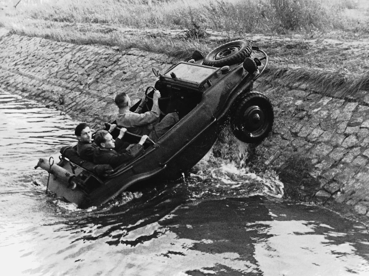 This image captures the defining versatility of the Type 166 Schwimmwagen as it transitions directly from water onto steep, uneven ground. Its watertight hull allowed it to cross rivers and flooded terrain, while four-wheel drive and aggressive gearing helped it regain traction once ashore. The rear-mounted propeller provided propulsion in the water, but the same compact Volkswagen drivetrain carried the vehicle forward on land. Few military vehicles of its size could move so seamlessly between both environments, making the Schwimmwagen one of the most adaptable machines derived from Porsche’s original small-car architecture.